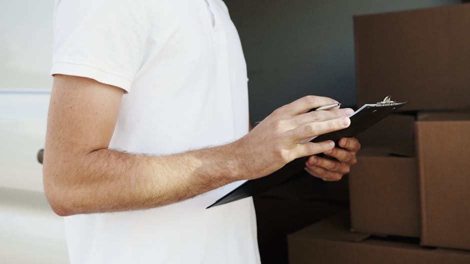 A person wearing a white short-sleeved shirt is holding a clipboard with one hand while the other hand uses a pen to take notes or check items. They are standing indoors in front of stacked cardboard 
