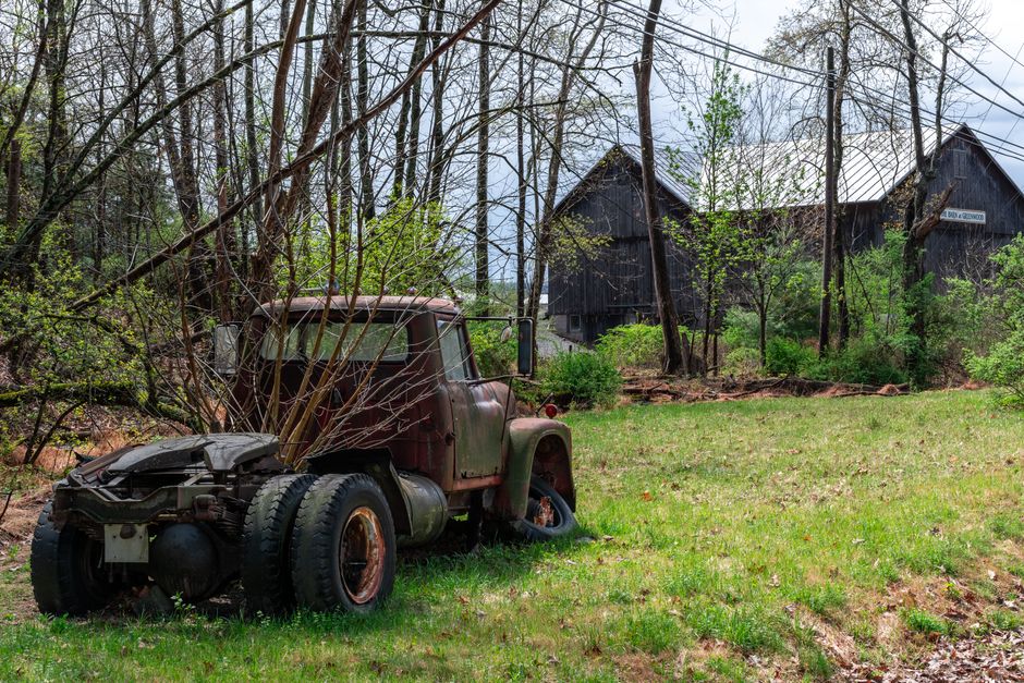 An old, rusted truck with a missing front wheel is situated on a grassy area in front of a wooded landscape, featuring bare trees and greenery. The truck's body shows signs of corrosion and age, with the hood slightly open and no visible driver or crew present. Behind the truck, there are wooden buildings or barns with dark, weathered exterior walls, one elevated on stilts. Overhead, power lines run across the scene, indicating proximity to residential or rural infrastructure. The setting suggests a rural environment, possibly during a house relocation or moving process where items are transported or stored temporarily. The image highlights the contrast between the dilapidated vehicle and the natural surroundings, relevant to logistics for home relocation, furniture transport, or packing and moving activities, as practiced by companies like Man with Van Barnes.