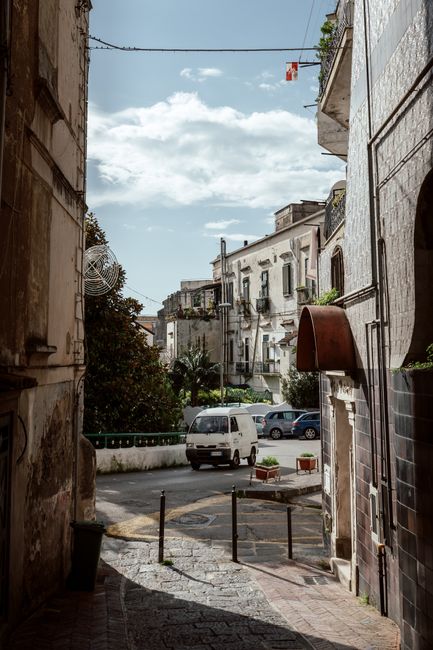 A narrow residential street in Castelnau with closely spaced multi-storey buildings featuring balconies, some with potted plants and iron railings. The street surface is paved with a combination of gr